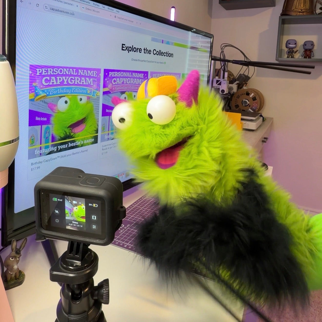 Green plush toy with a camera on a tripod in front of a computer screen displaying a website.